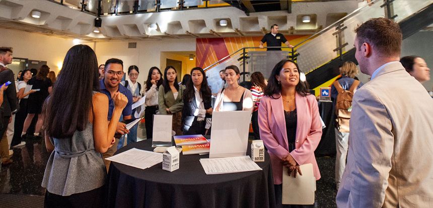 Students and employers talk around a table at a busy campus career fair, with groups of attendees gathered throughout a large indoor hall.