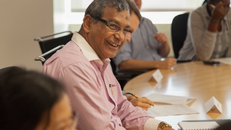 A man wearing a pink shirt and glasses is caught mid-laugh as he attends a meeting in a classroom.