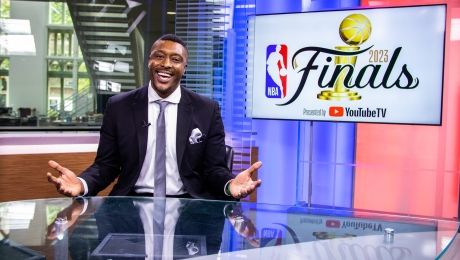 An NBA athlete sits at a broadcast table in a USC Annenberg newsroom, smiling and holding his hands out to camera. The screen next to him has the NBA and YouTube logos on it, and says “2023 Finals.”