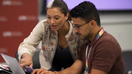 An instructor leans over a student’s computer, pointing at the screen.