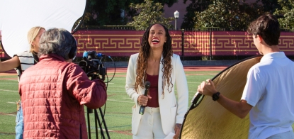 An NBA athlete sits at a broadcast table in a USC Annenberg newsroom, smiling and holding his hands out to camera. The screen next to him has the NBA and YouTube logos on it, and says “2023 Finals.”