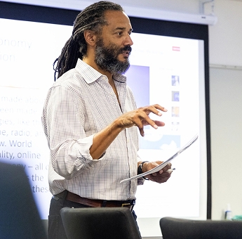 Professor Ben Carrington stands at the head of a classroom, giving instruction mid-sentence, in front of a projected screen and holding a piece of paper.